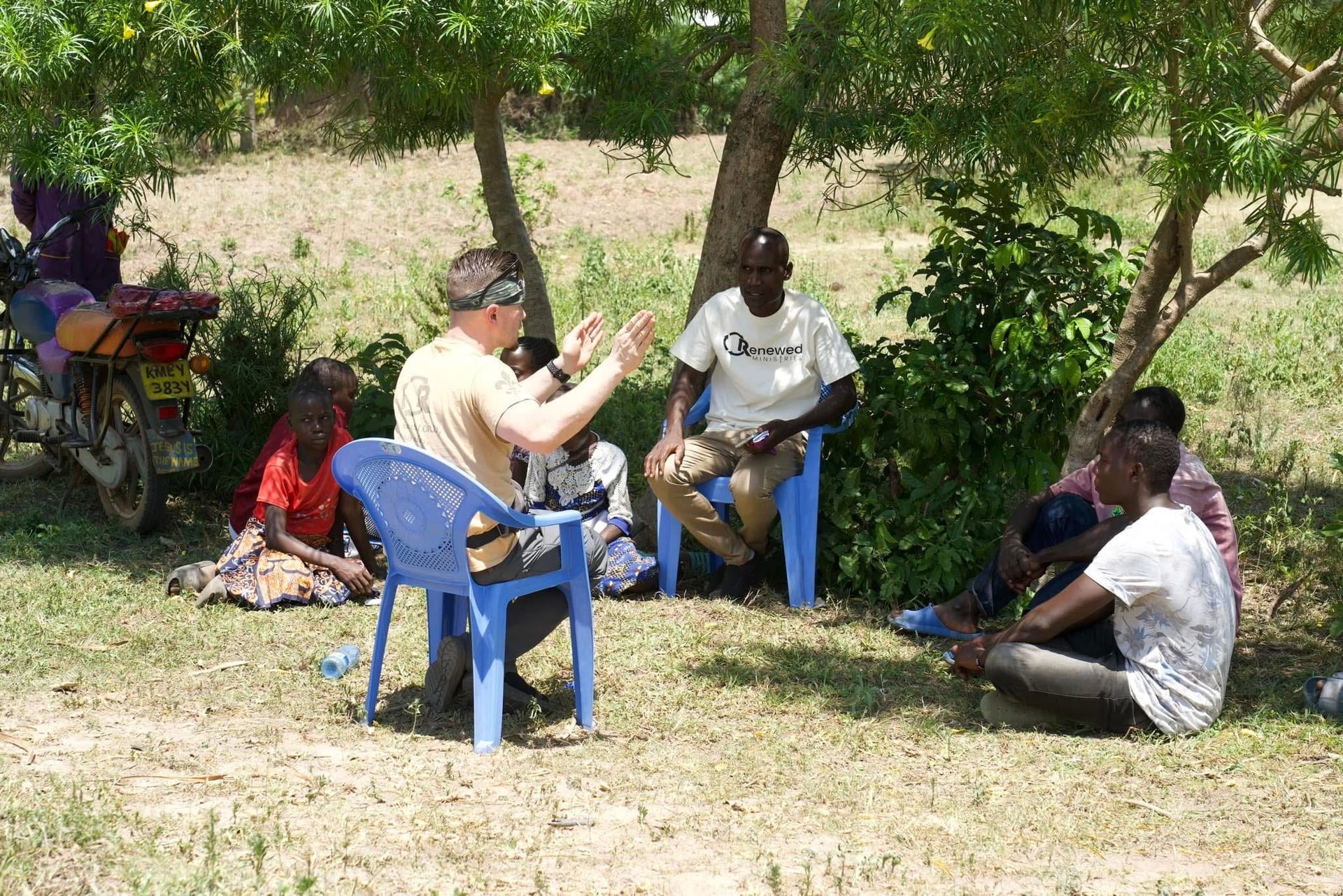 Group of people seated outdoors, listening to a person gesturing. A motorcycle is parked nearby, under a tree.