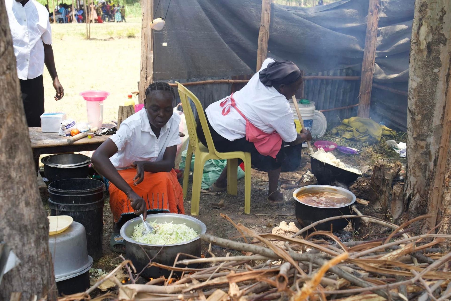 Two people cooking outdoors over a fire. One stirs a pot, the other sits.
