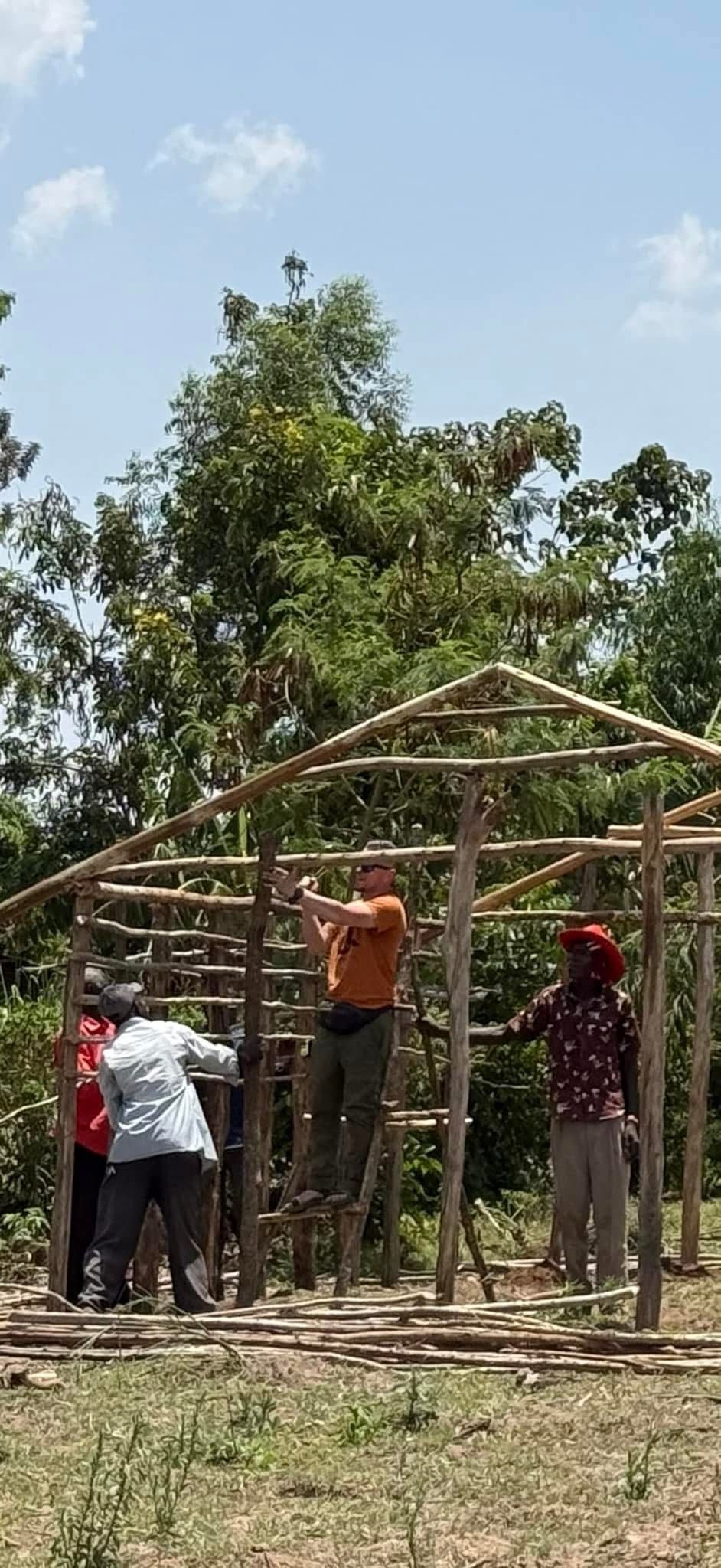 People constructing a wooden structure outdoors. They work on a frame of a roof, green trees in background.