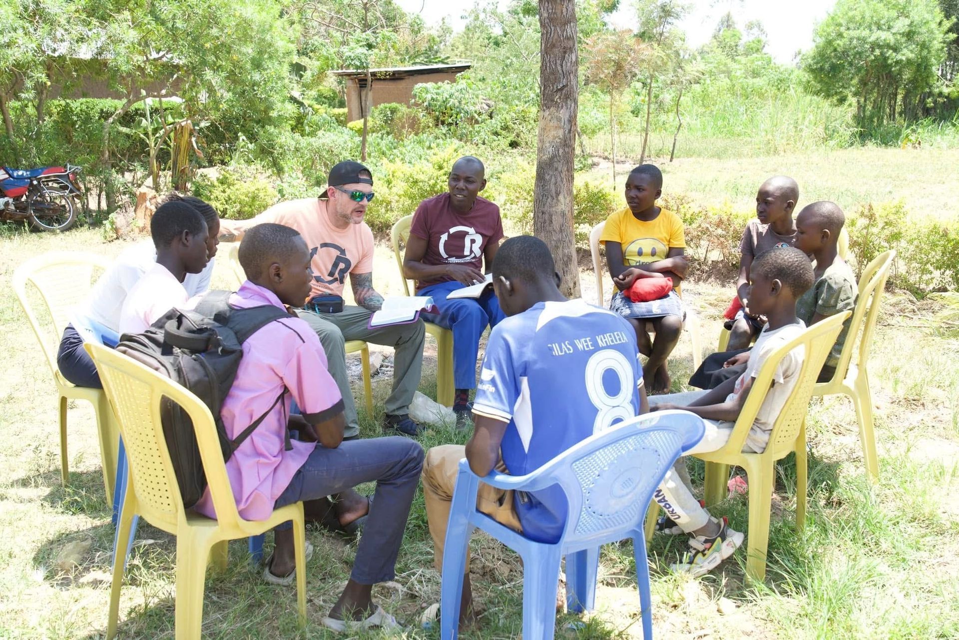Group of people seated in a circle outdoors, talking. A man is holding papers. Yellow and blue chairs. Sunny day.