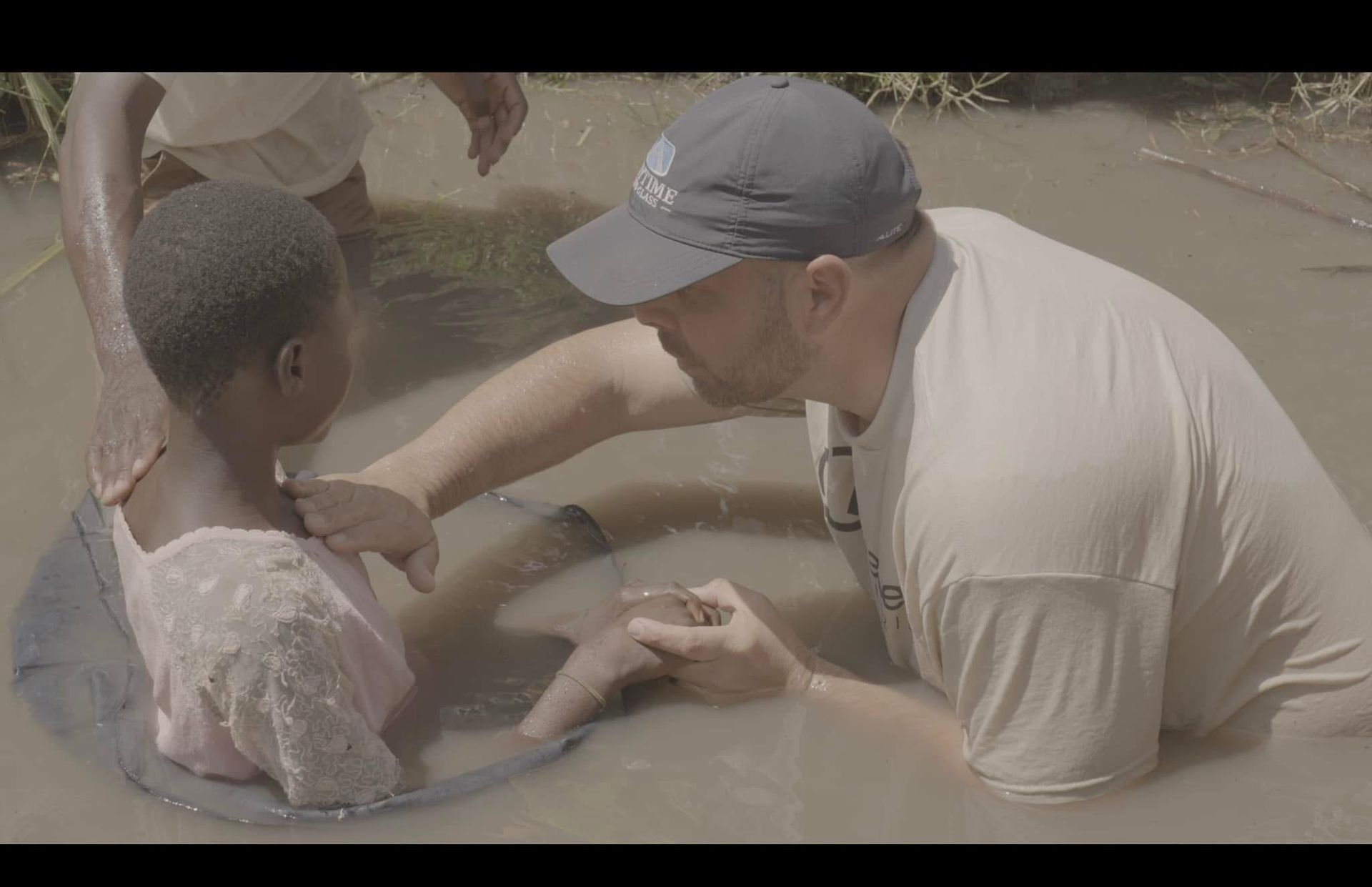 Man baptizes a child in murky water; both are partially submerged.