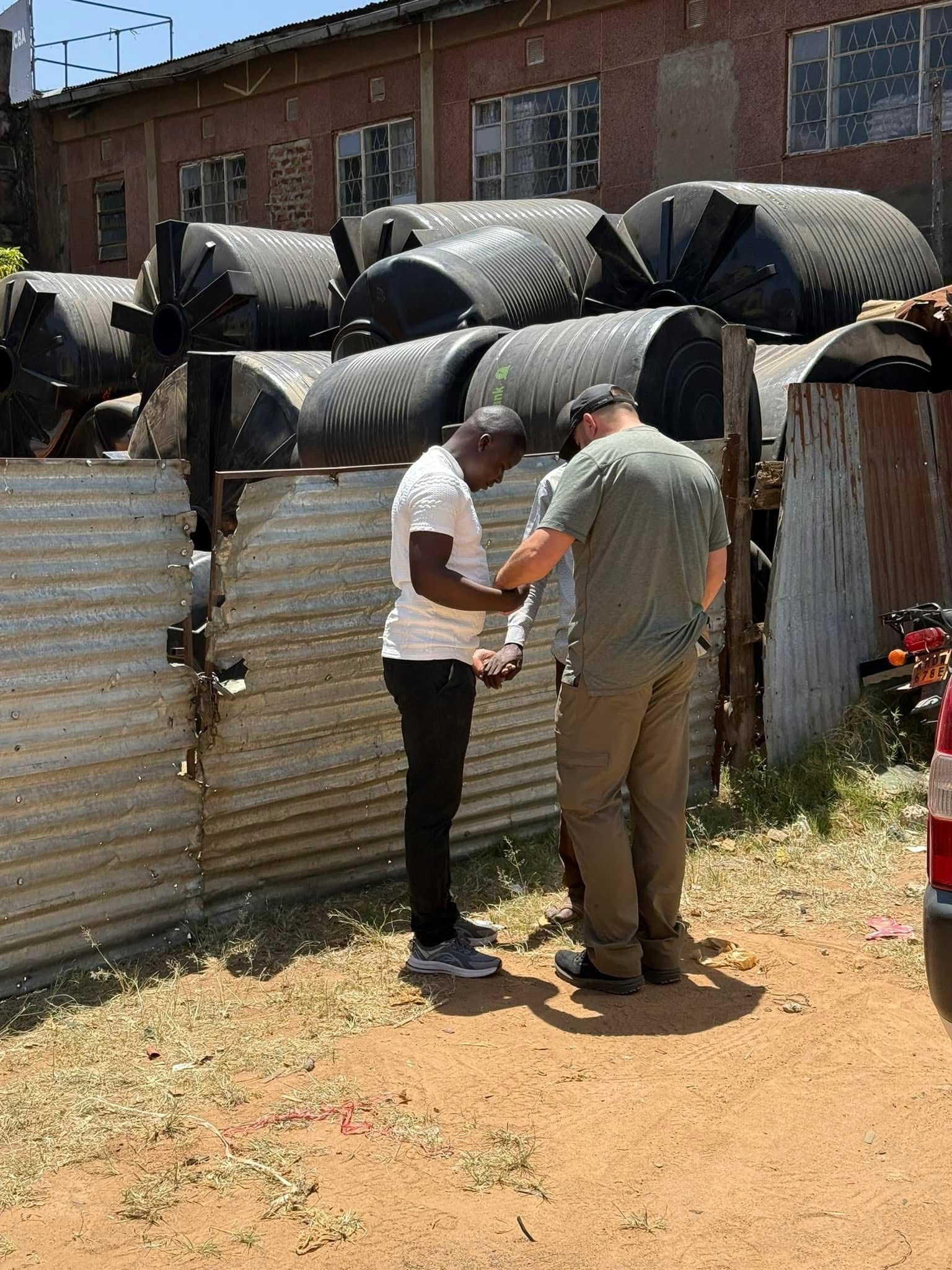 Two men standing near a corrugated metal fence, looking at something. Black water tanks in the background.
