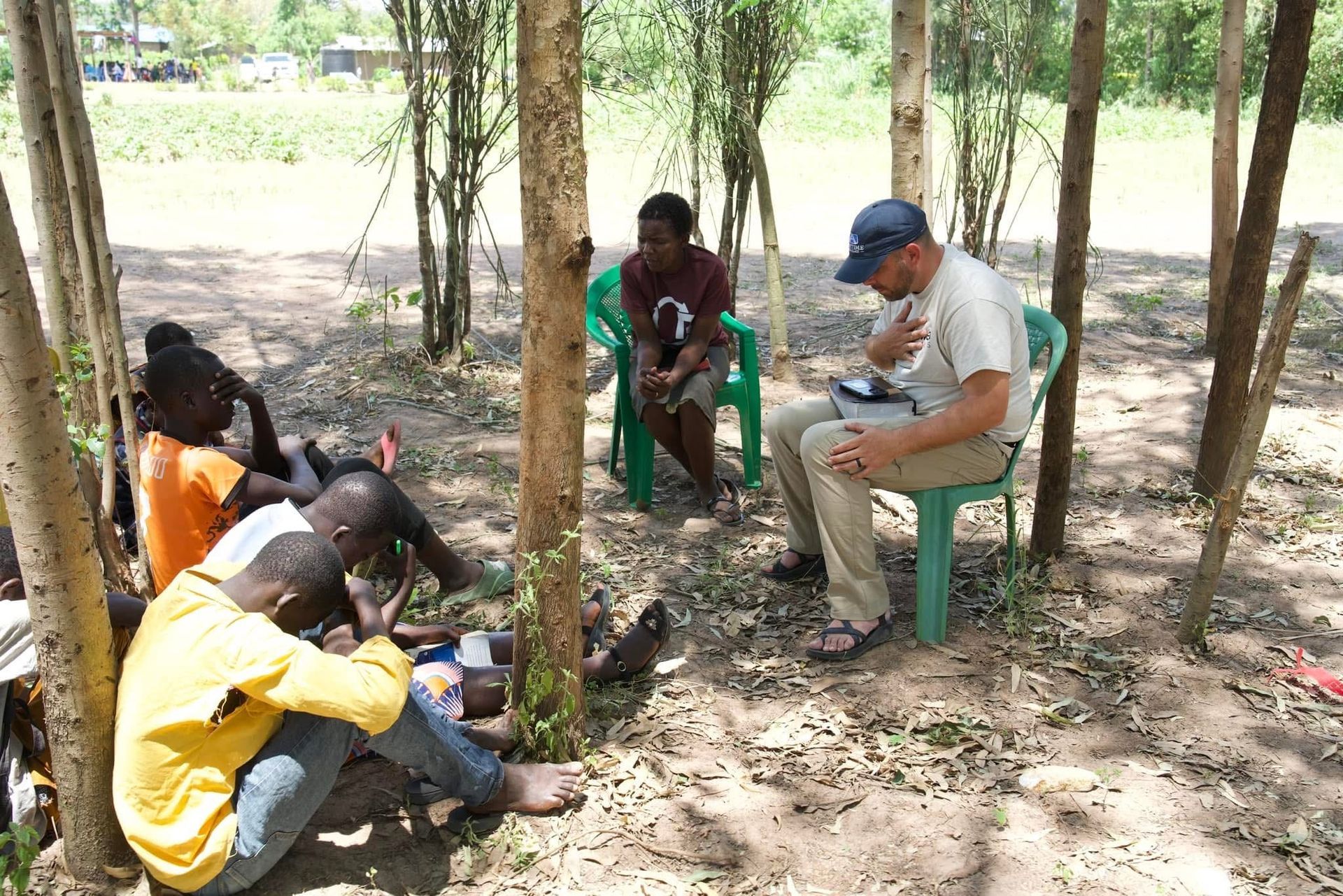 A man in a hat talking to people seated outdoors under trees.