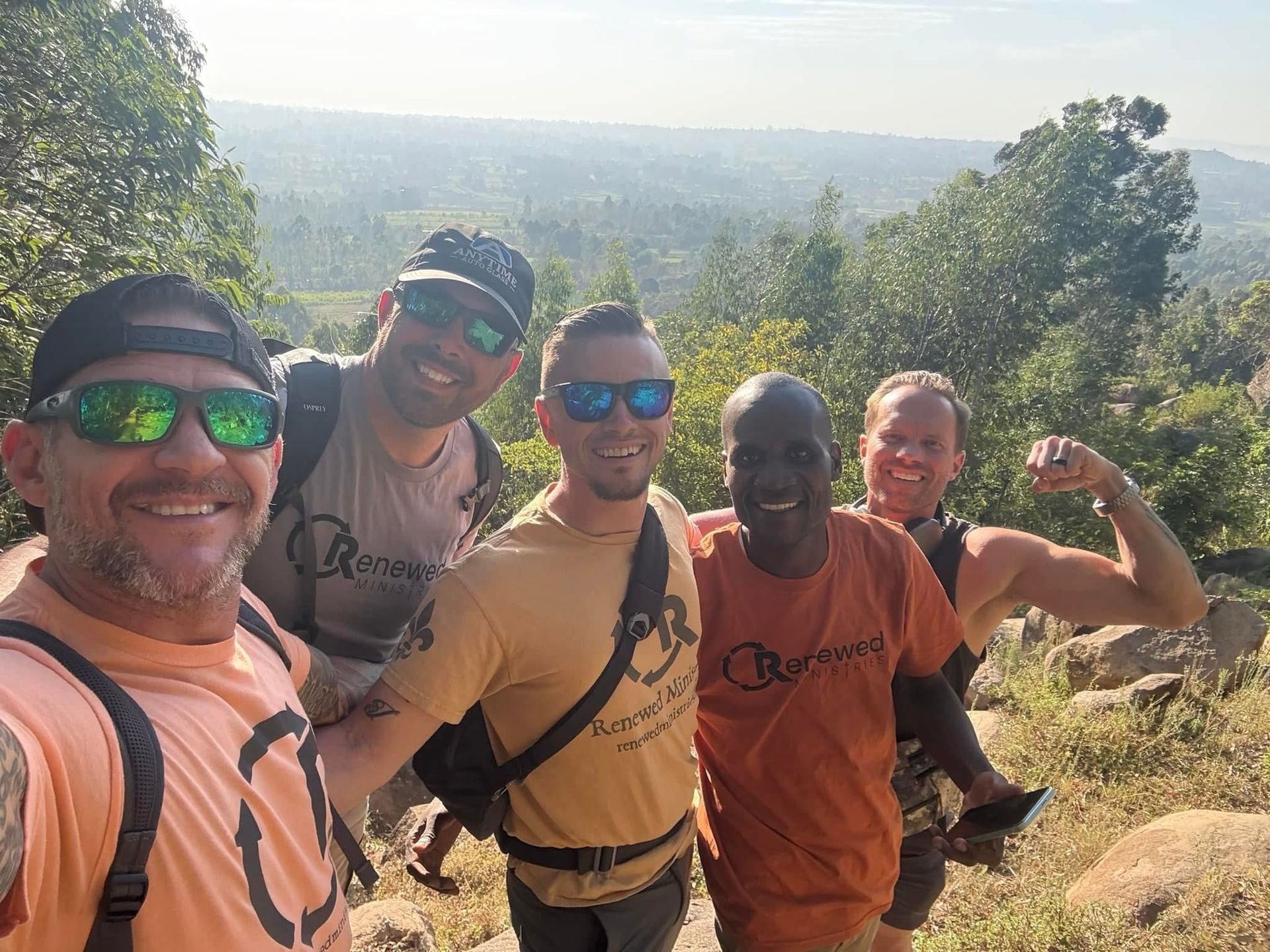 Five people in sunglasses pose smiling outdoors with a mountain view.