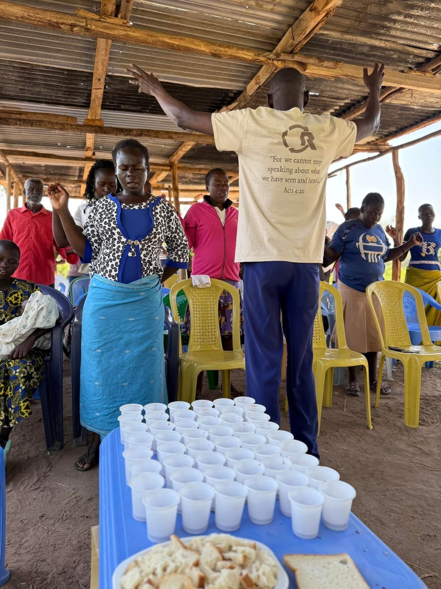 People in a gathering with raised hands, possibly in prayer. A table with cups and bread in front.