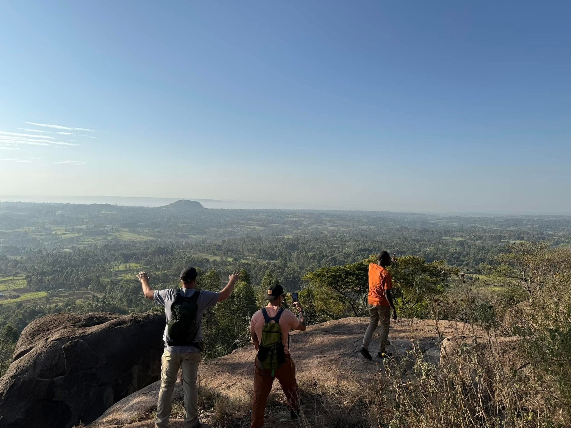 Three hikers atop a rocky hill with arms raised, overlooking a green valley under a blue sky.