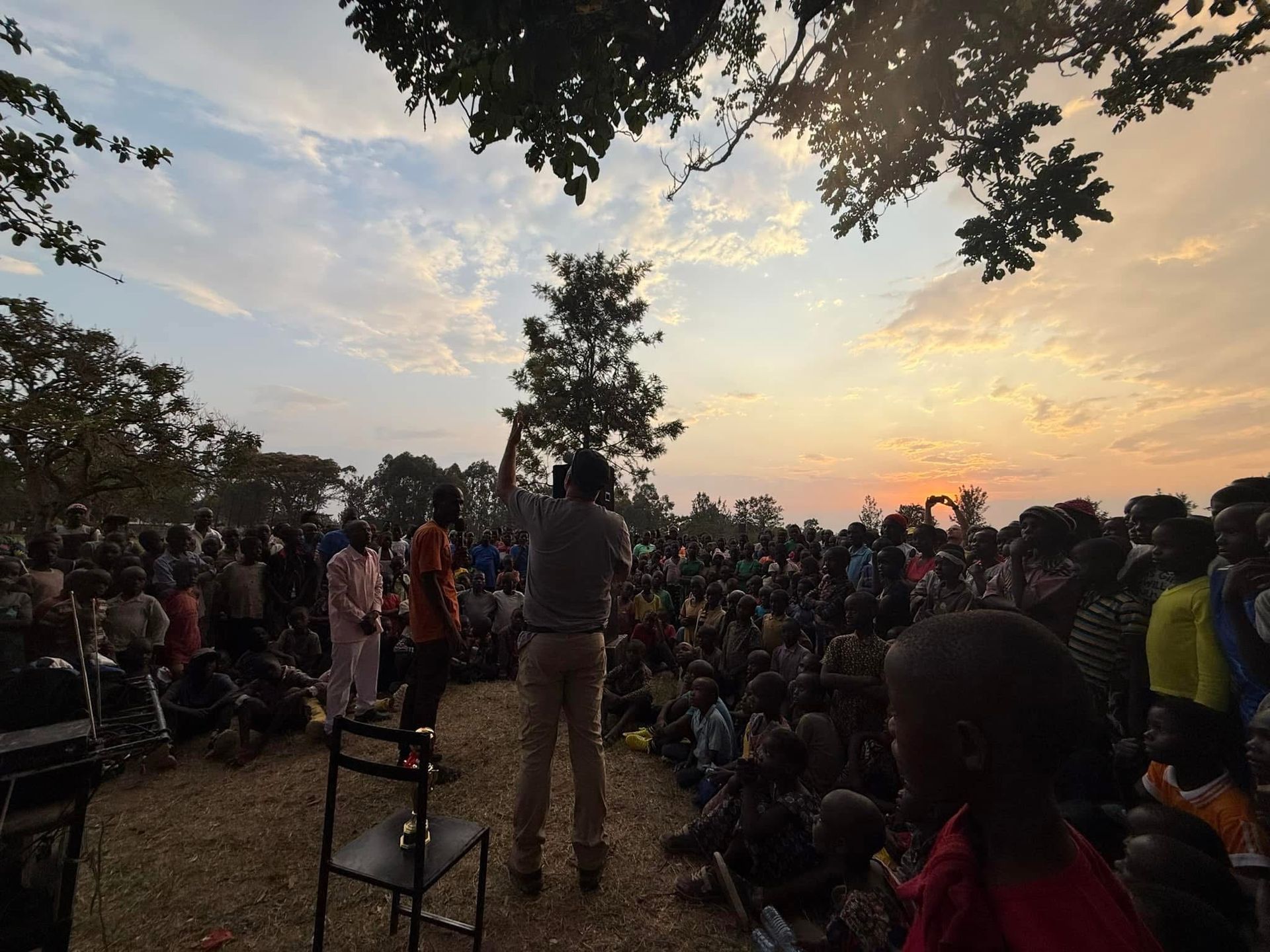 Large crowd gathered outdoors at sunset; a person on stage appears to be speaking or performing.