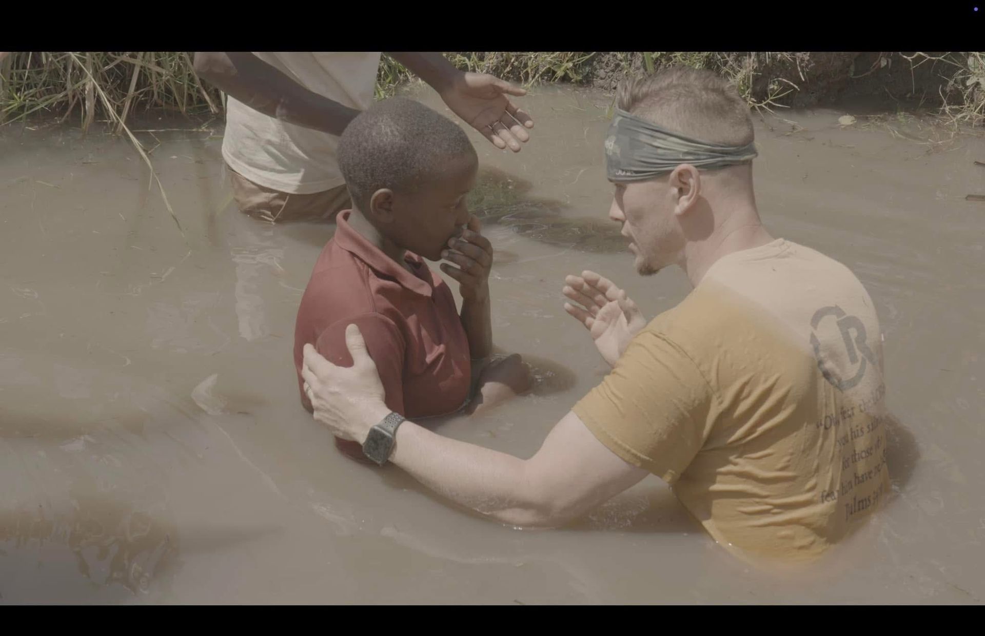 Man baptizes child in murky river, two others watch.