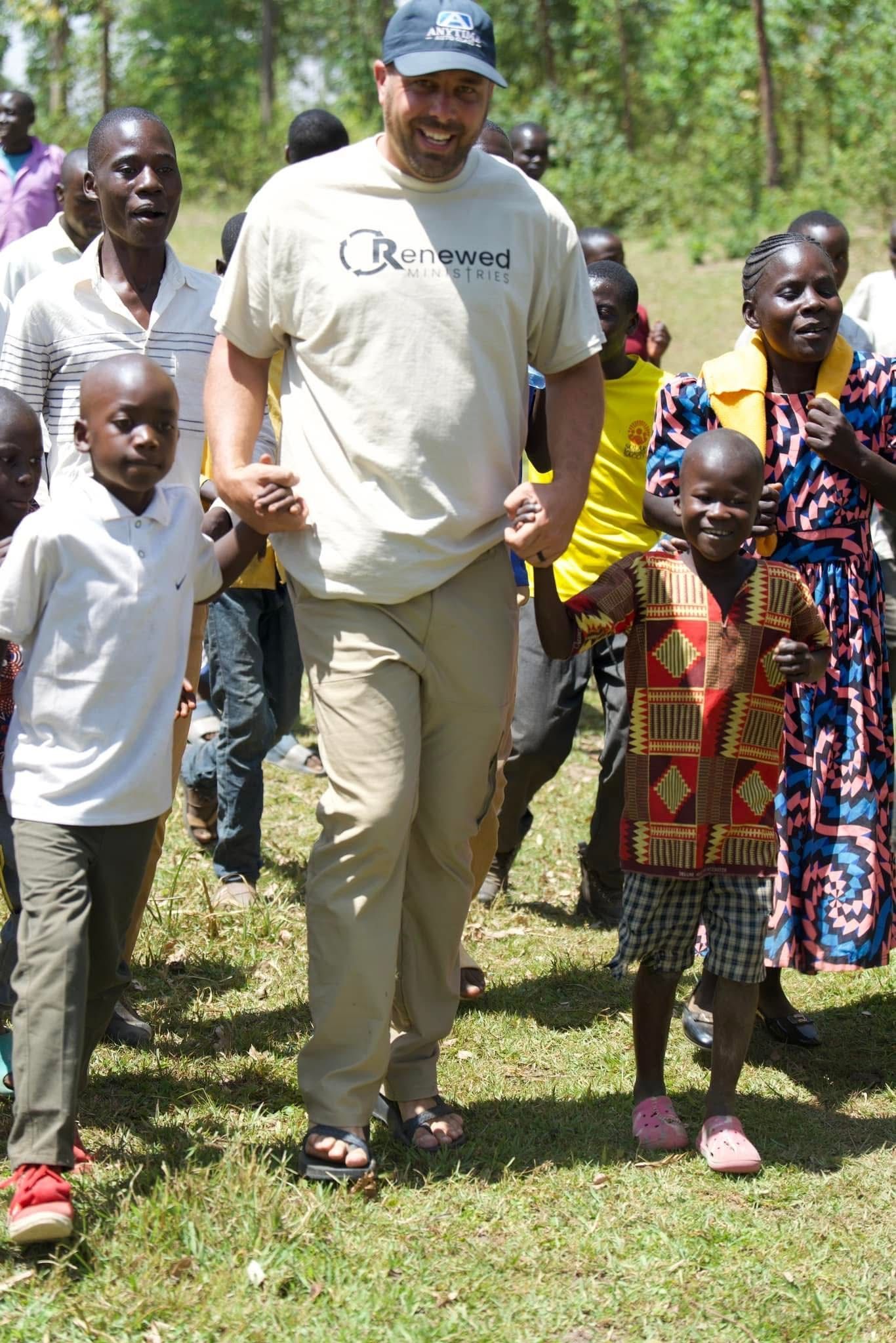 Man in tan shirt and khaki pants walks with group of children in an outdoor setting.