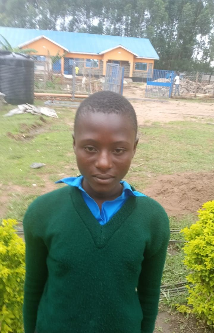 Young child with shaved head, wearing patterned blue and floral top, looking directly at the camera, outdoors.