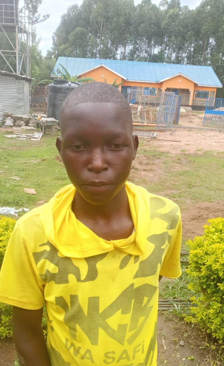 A young Black person stands in front of blue bars. They wear a white shirt with a green and black jacket.