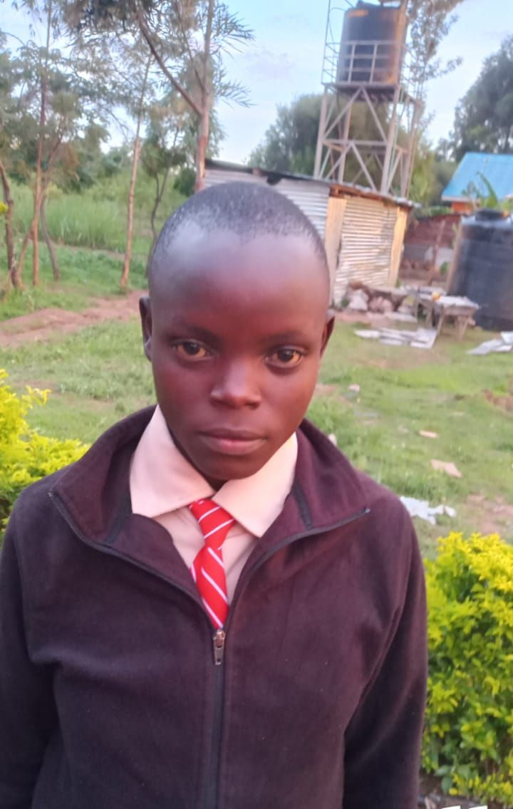 A young Black person stands in front of blue bars. They wear a white shirt with a green and black jacket.