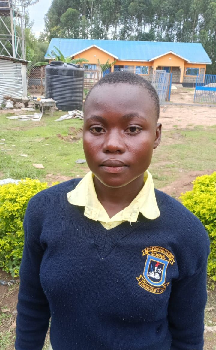 A young Black person stands in front of blue bars. They wear a white shirt with a green and black jacket.