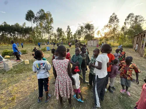 Children gathered outdoors, looking up. Trees and a wooden structure are in the background. Bright sunlight.