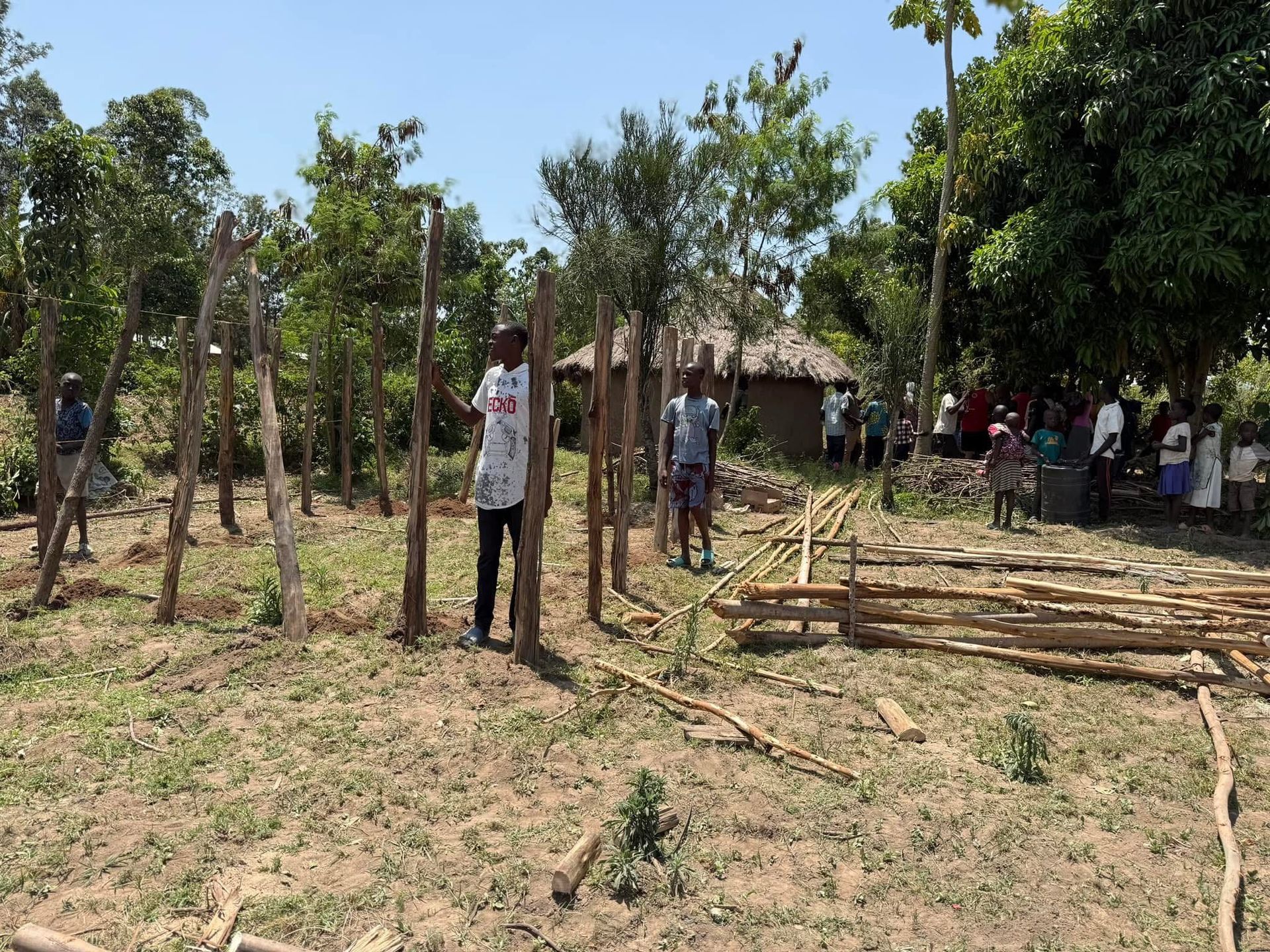 People building a structure with wooden posts in a field; small hut and trees in background.