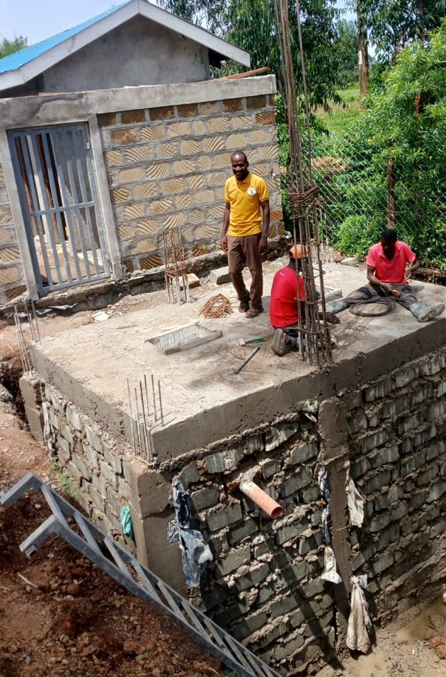 Construction of a single-story building. Stone walls are in progress, set amidst trees and greenery.