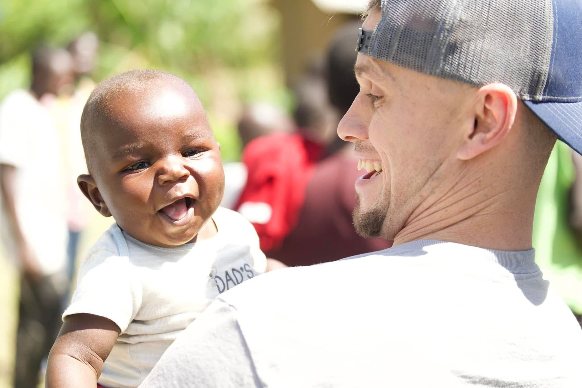 Man holding a smiling baby, both looking at the camera. Baby wears a white shirt, man wears a cap.