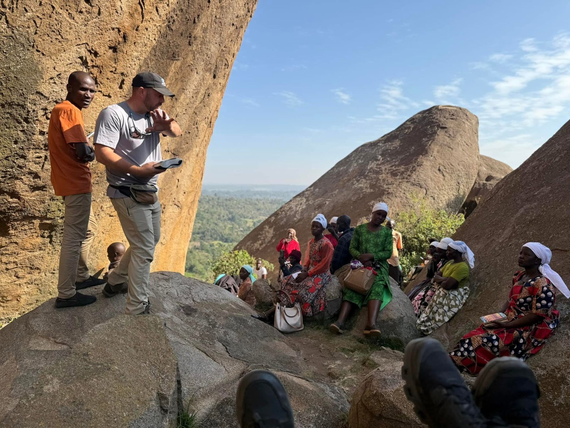 People gathered on rocks with a man speaking, landscape visible.