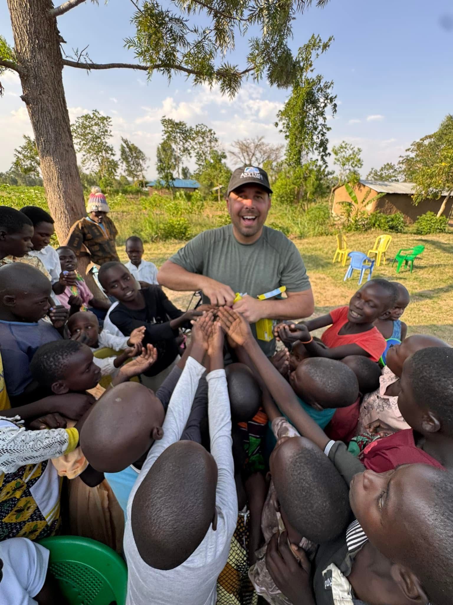 Man surrounded by children, reaching up. They are outdoors near trees and buildings.