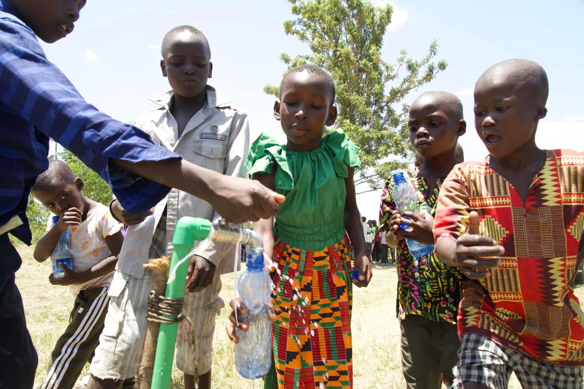 Children at an outdoor water pump, filling plastic bottles.