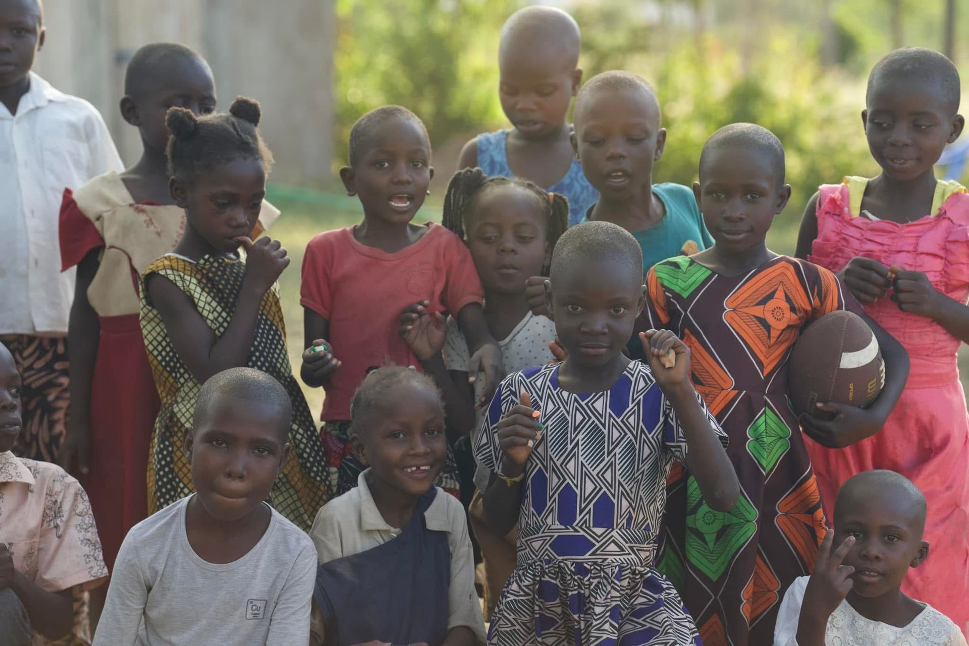 Group of children smiling outdoors, some holding a football, others in colorful clothing.