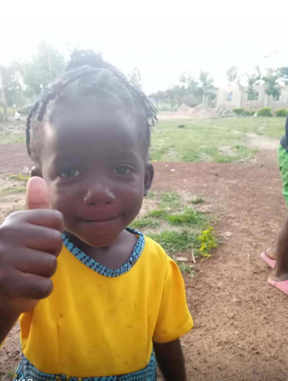 Smiling Black child giving a thumbs up, wearing a yellow shirt, outdoors on a sunny day.