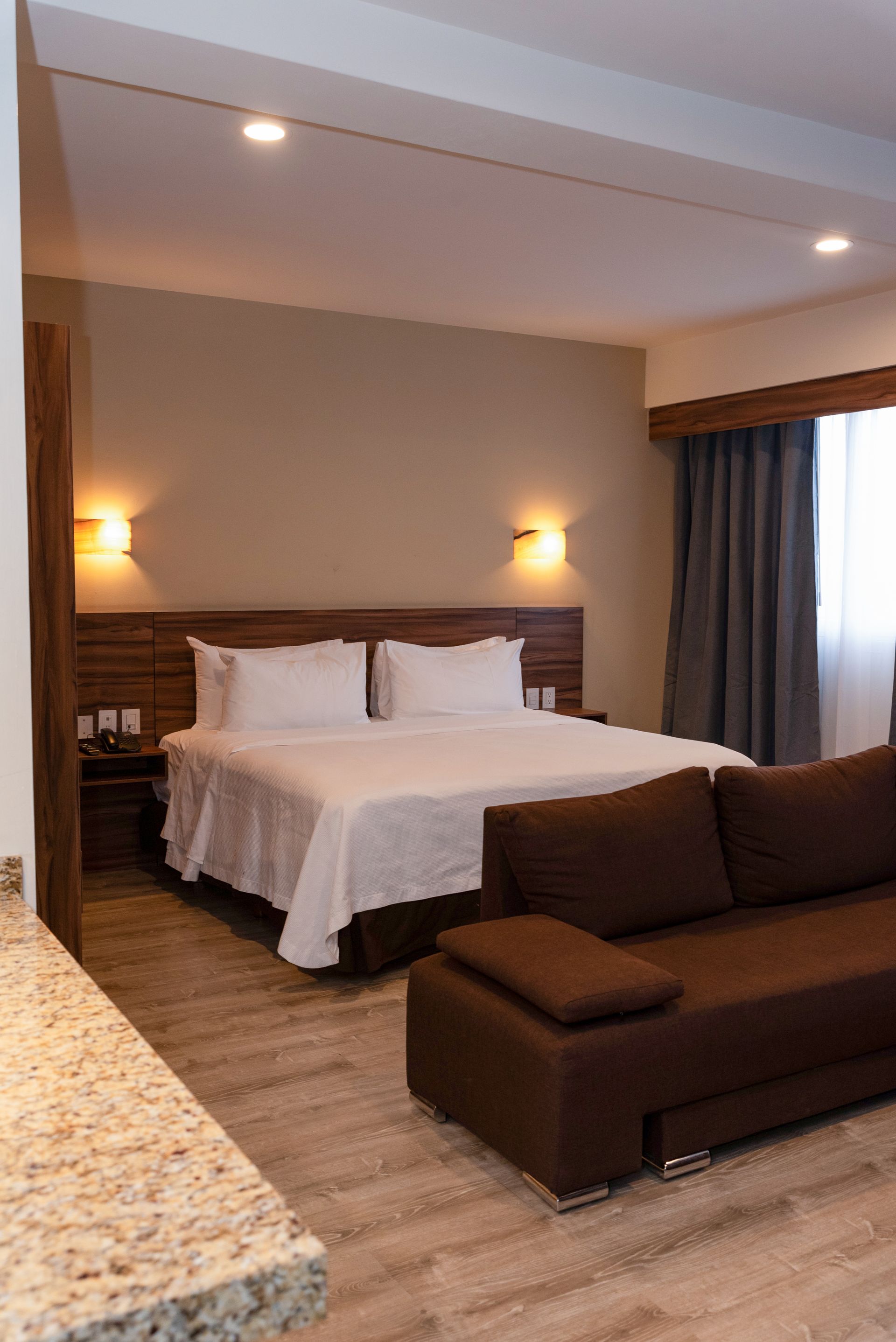 Habitación de hotel con cama, sofá y encimera de granito. Tonos marrones y beige.
