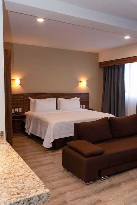 Habitación de hotel con cama, sofá y encimera de granito. Tonos marrones y beige.