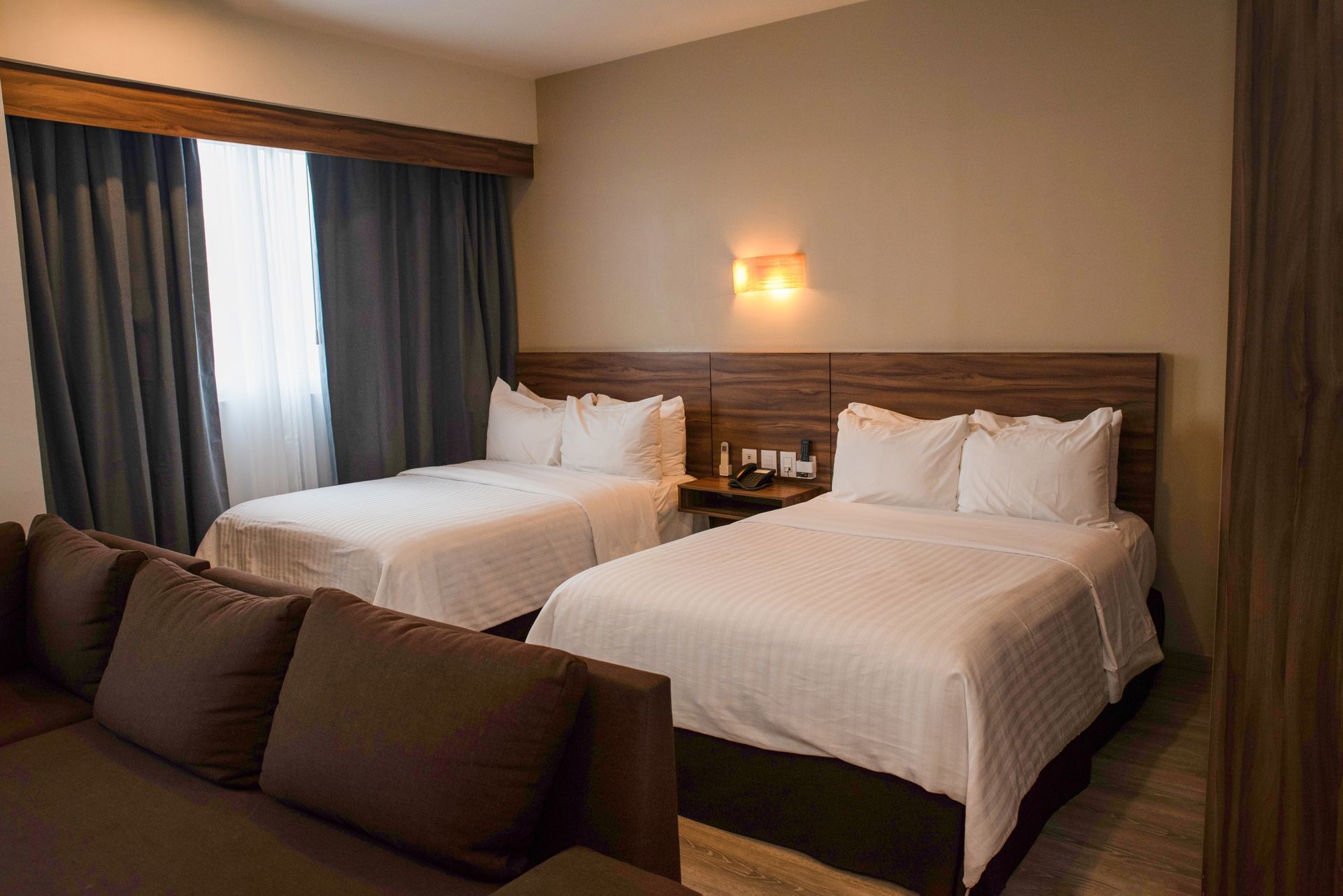 Habitación de hotel con dos camas dobles, ventana, sofá y mesita de noche con lámpara.