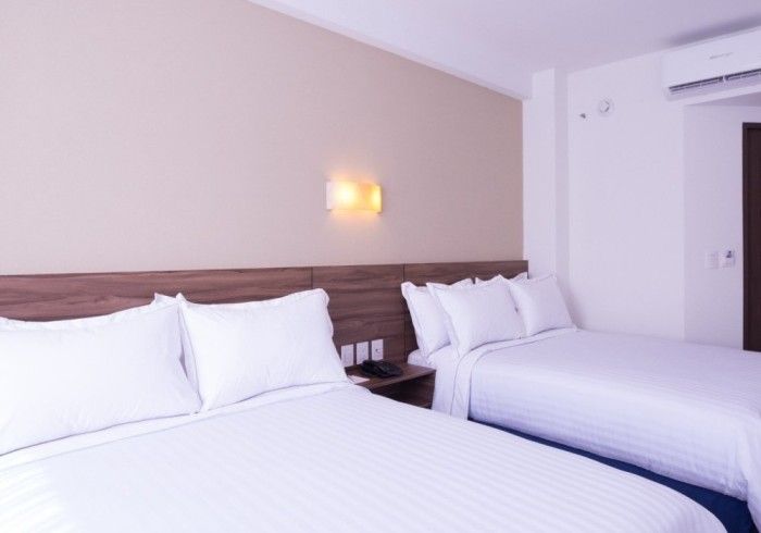 Habitación de hotel con dos camas cubiertas con sábanas y almohadas blancas, paredes en tonos neutros y aire acondicionado.