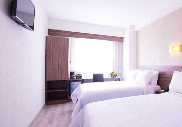 Habitación de hotel con dos camas individuales, un pequeño escritorio, armario y una ventana con luz natural.