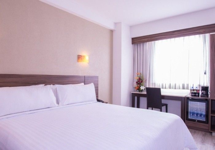 Habitación de hotel con cama, escritorio, mininevera y ventana con cortinas; colores neutros.