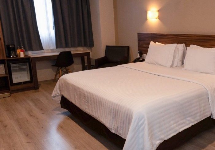 Habitación de hotel con cama, escritorio, silla, mininevera y ventana con cortinas oscuras.