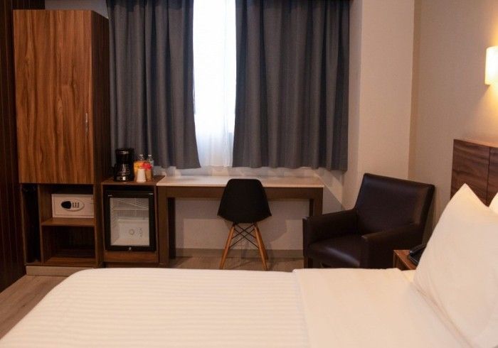 Habitación de hotel con cama, escritorio, silla, armario, mininevera y ventana con cortinas.