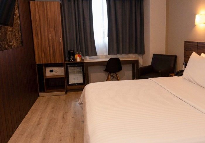 Habitación de hotel con cama, armario de madera con caja fuerte y mininevera, escritorio y sillón.