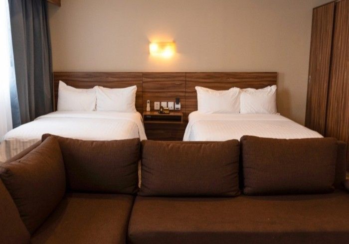 Habitación de hotel con dos camas, sofá marrón, paredes en tonos neutros, cabeceros de madera y mesita de noche.