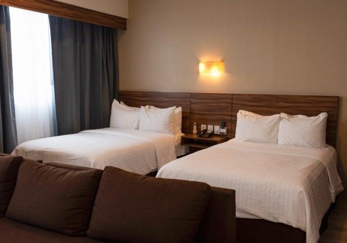 Habitación de hotel con dos camas, sofá marrón y ventana con cortinas grises.