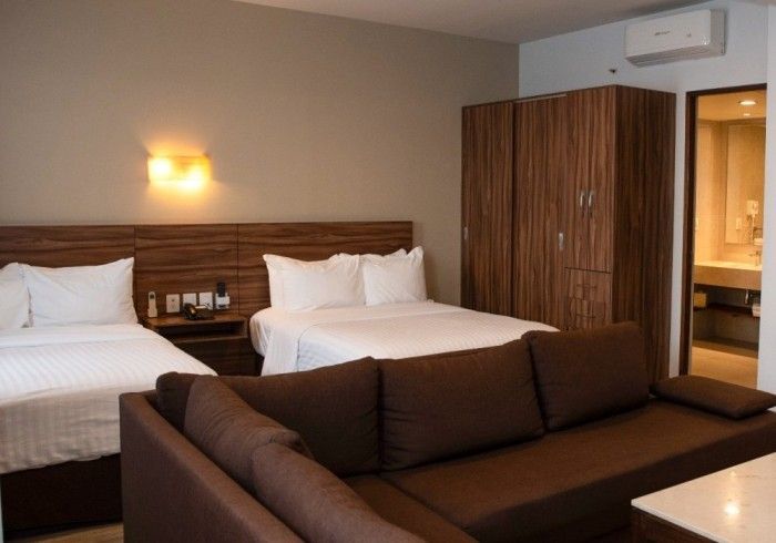 Habitación de hotel con dos camas, sofá marrón y armario, con baño a la vista.