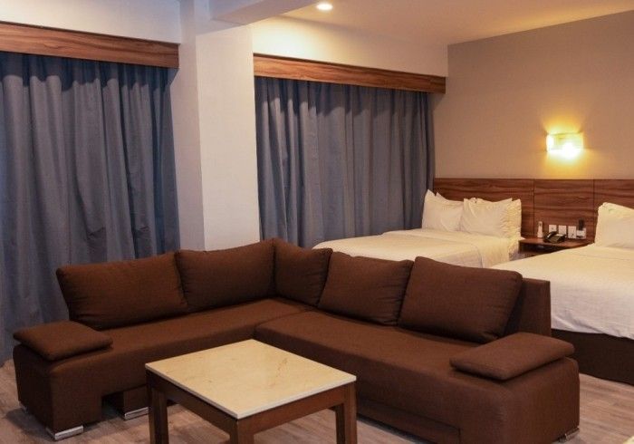Habitación de hotel con sofá seccional marrón, dos camas y cortinas azules.
