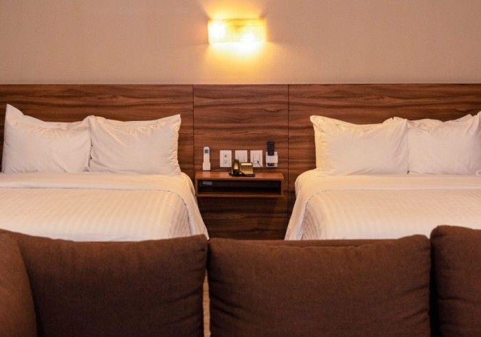 Dos camas tamaño queen con sábanas blancas, una mesita de noche y un cabecero en tonos cálidos. Cojines marrones en el sofá, en primer plano.