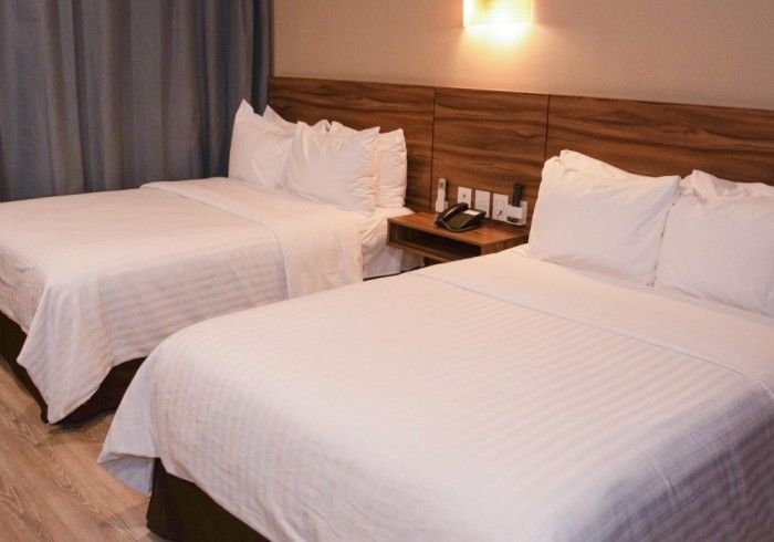 Dos camas con ropa de cama blanca en una habitación de hotel, mesita de noche pequeña, cabecero de madera.