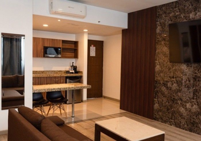 Moderna suite de hotel con cocina americana, muebles marrones y televisor montado en una pared texturizada.