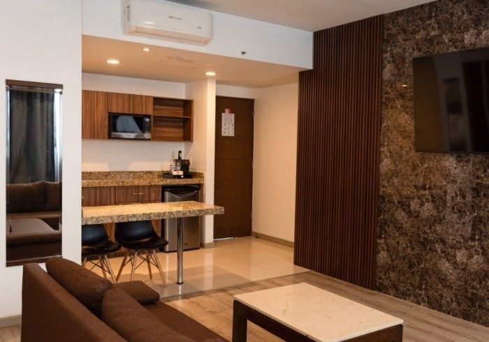 Una habitación de hotel moderna con cocina americana, sofá marrón, televisor y mesa de comedor pequeña.
