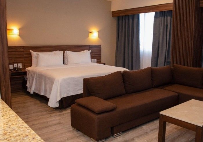 Habitación de hotel con cama, sofá seccional marrón, mesita y ventana con cortinas.