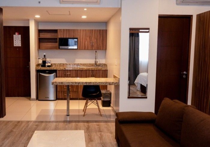 Habitación de hotel pequeña con cocina americana, sofá marrón y una mesa con una silla negra.