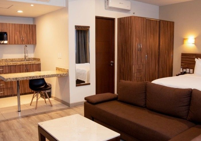 Suite de hotel con cocina americana, zona de estar y dormitorio. Sofá marrón, armarios de madera y encimeras de granito.