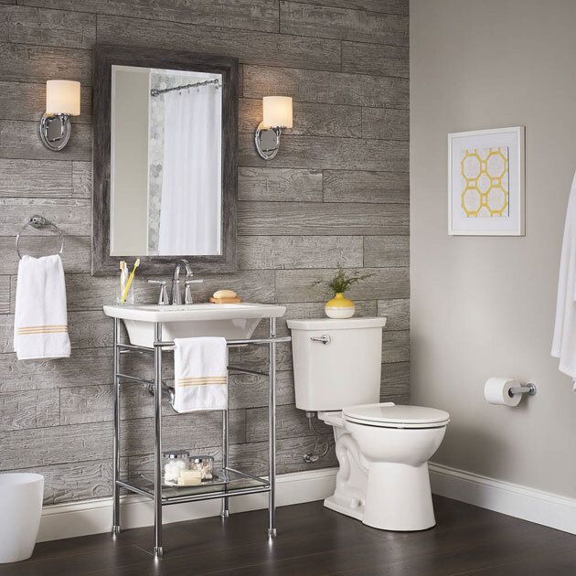 Bathroom with gray wood-look wall, pedestal sink, toilet, mirror, towel rack, wall sconces, artwork, and dark floor.