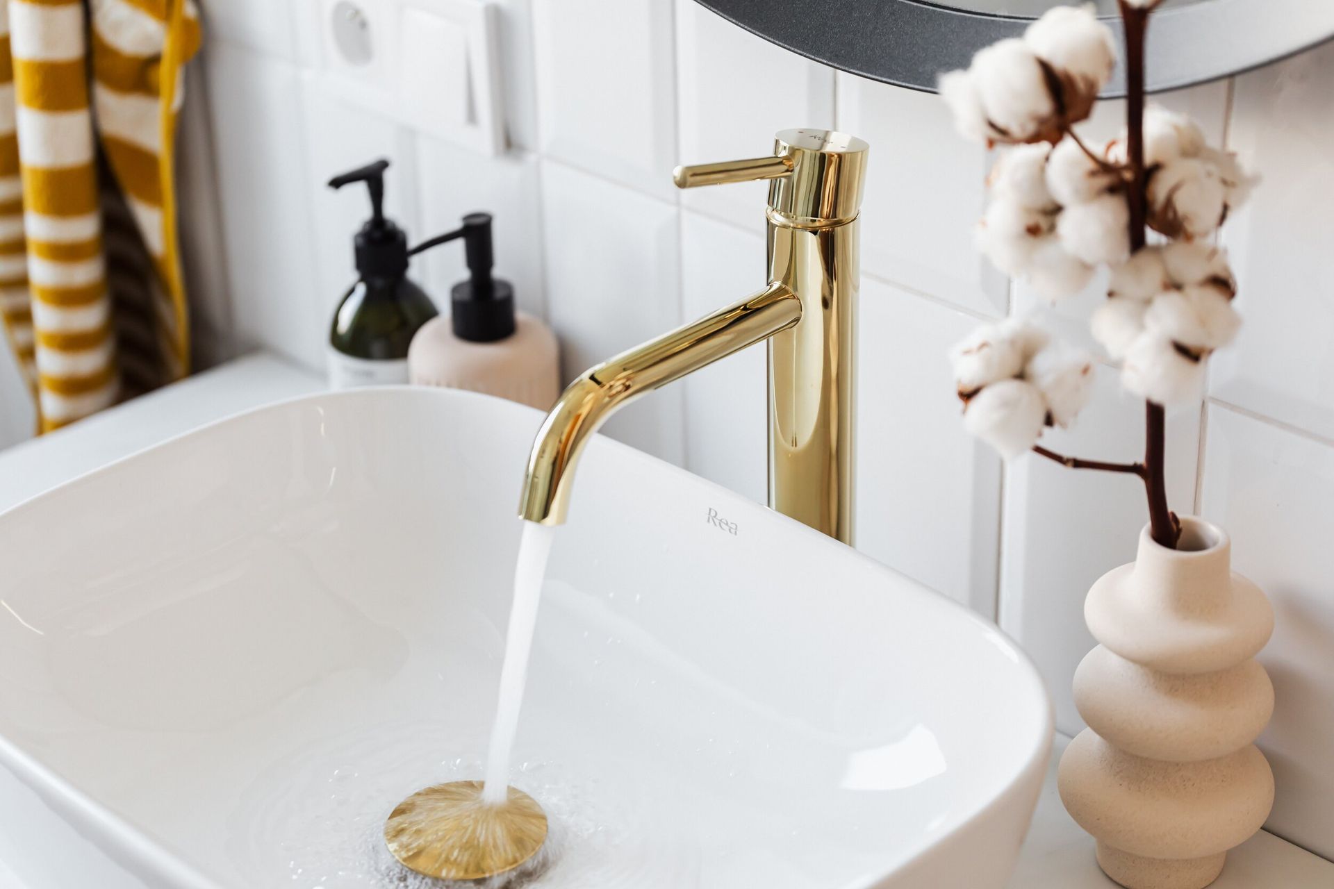 Gold faucet with water flowing into a white bathroom sink, with a vase of cotton and soap dispensers.