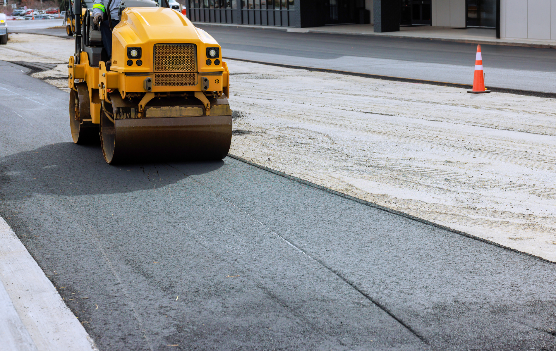 Yellow road roller compacting fresh asphalt on a road, with a partially paved section beside it.