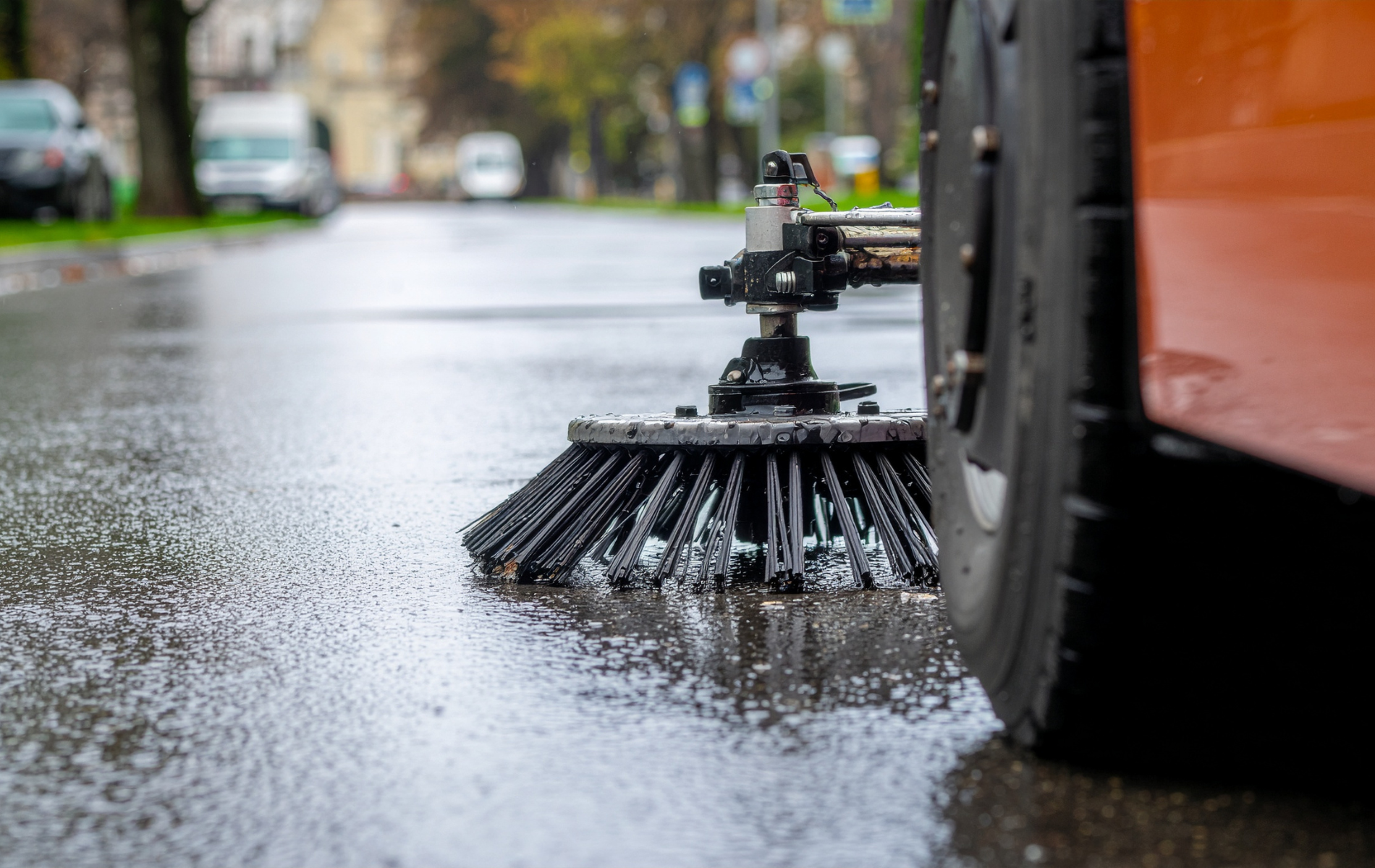 Street sweeper scrubbing a wet road, orange vehicle details visible.